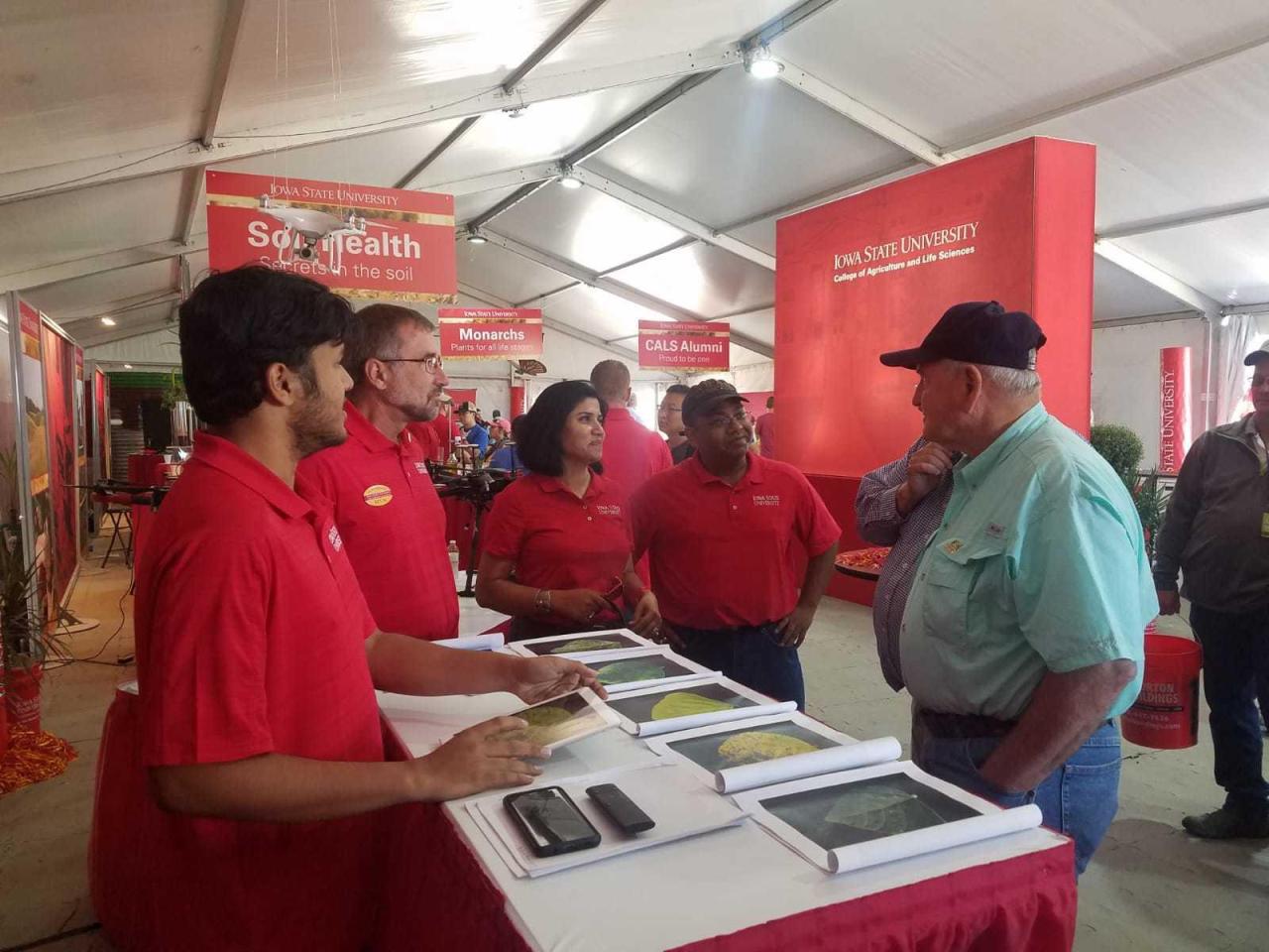 Iowa State team in red shirts at a table of soybean leaf images, talking with a visitor in a cap inside an exhibit tent.