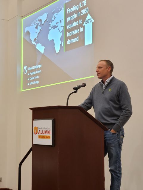 Speaker at an ISU Alumni Center podium in front of a slide on feeding 9.7 billion people by 2050.
