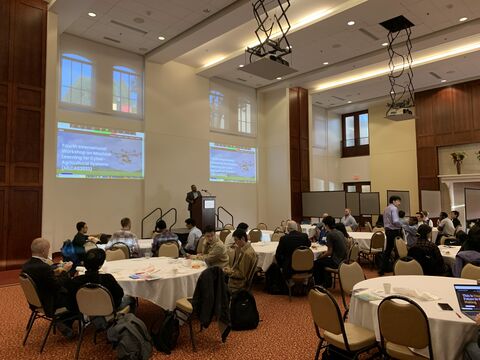 Ballroom with attendees at round tables facing a speaker and two screens showing the workshop title.