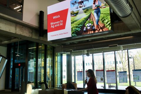 Presenter with a microphone below a large screen reading "WIAA Women in Ag and AI" in a glass-walled atrium.