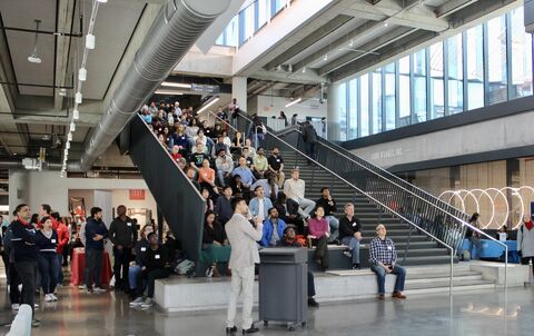 Audience seated on a large indoor staircase listening to a speaker in a modern atrium.