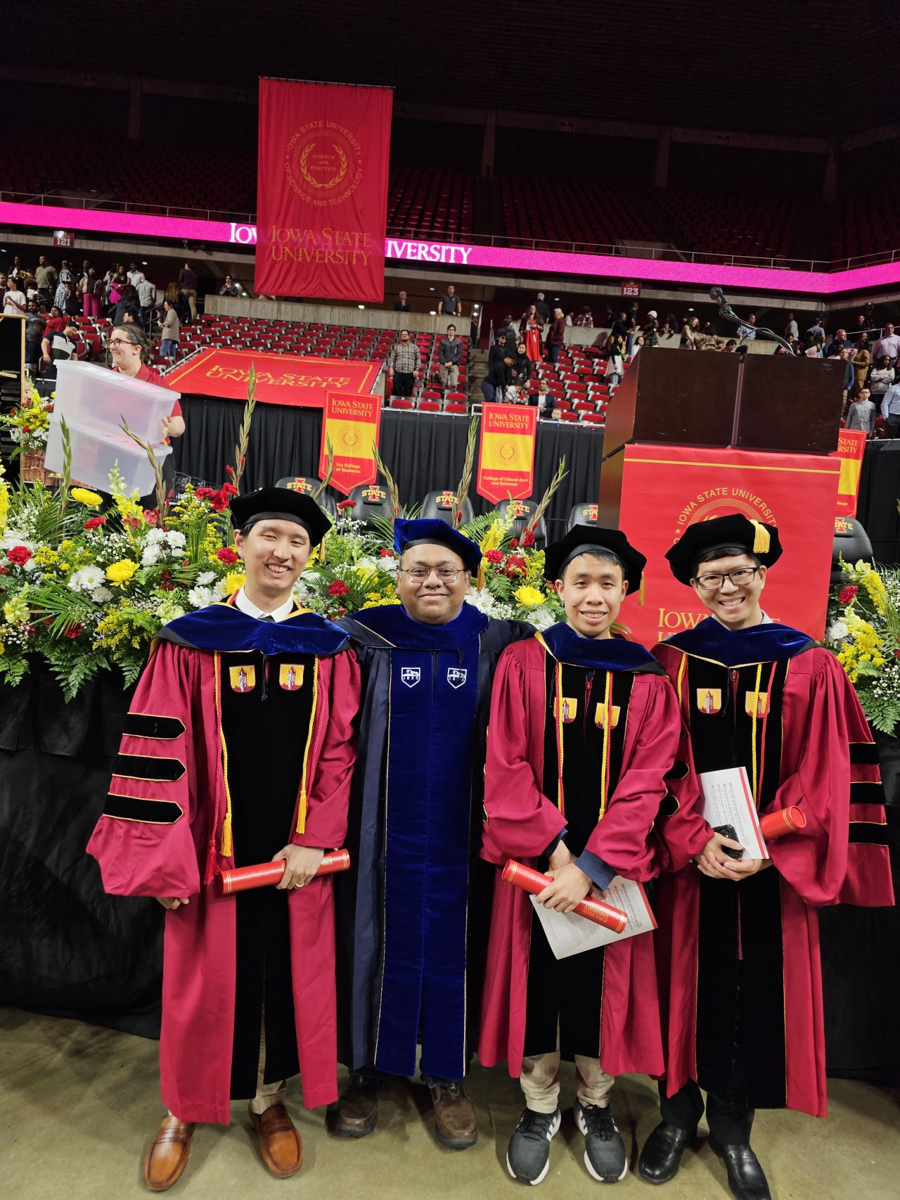 Three new doctoral graduates in cardinal gowns and their advisor in blue regalia in front of flowers and Iowa State banners.