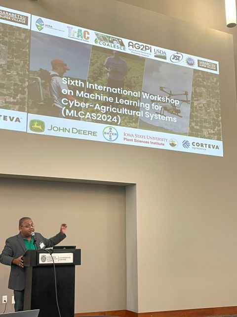 Speaker at a podium beneath a large screen titled "Sixth International Workshop on Machine Learning for Cyber-Agricultural Systems (MLCAS2024)" with sponsor logos.