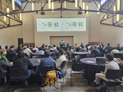 Wide view of a full conference hall with attendees at round tables facing a presenter and twin screens.