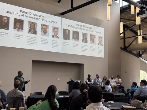 Panelists seated at the front of a hall below a projected slide listing panel members with photos.