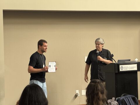 Young man holding a certificate beside a presenter speaking into a microphone at a podium.