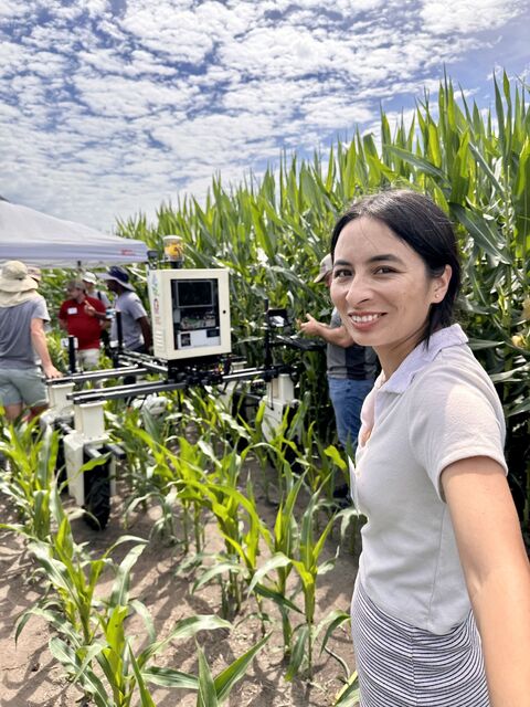 Woman smiling in a cornfield next to a wheeled field robot carrying a sensor box, with people under a tent behind.