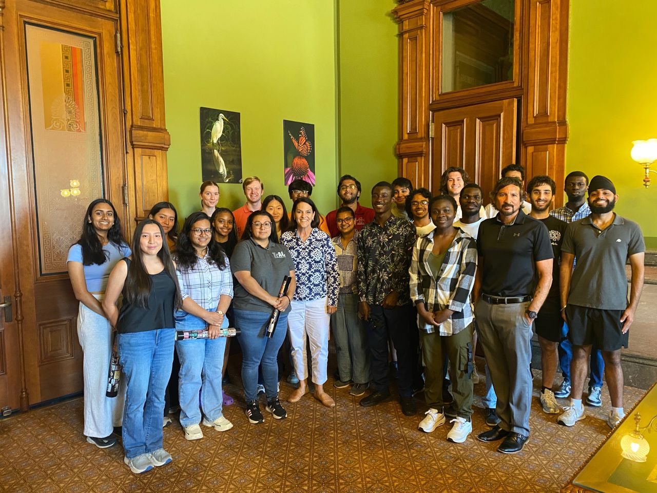 Large group of about twenty-five students and staff posing in an ornate room with wood-paneled doors and green walls.