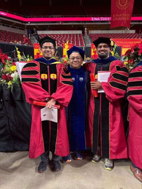 Two new doctoral graduates in cardinal Iowa State gowns standing with their advisor in blue academic regalia in an arena.