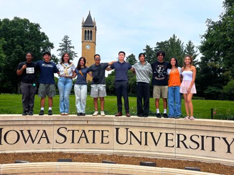 Eleven young people standing on the curved Iowa State University stone sign, with the Campanile clock tower and trees behind.
