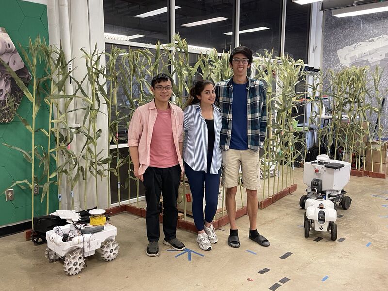 Three people standing between two small white wheeled robots in front of an indoor stand of tall corn plants.