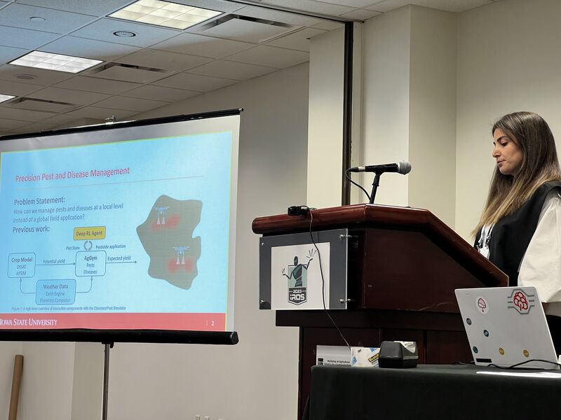 A presenter at an IROS 2023 podium next to a projected slide titled "Precision Pest and Disease Management".