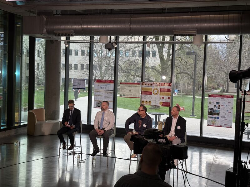 Four panelists seated on stools in a glass-walled atrium with research posters on the windows behind them.