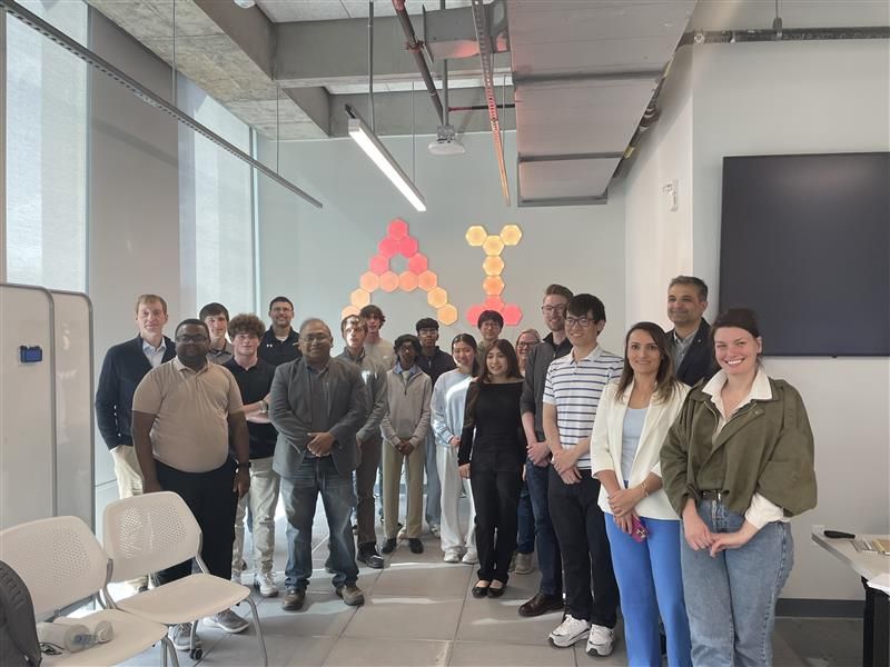 About twenty-five people posing for a group photo in a bright room with hexagonal light panels on the wall.