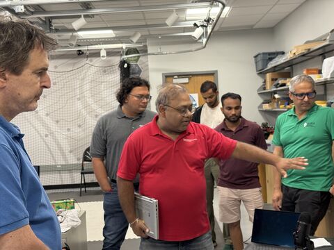 A man in a red shirt gestures toward a screen while several visitors watch in a robotics lab.