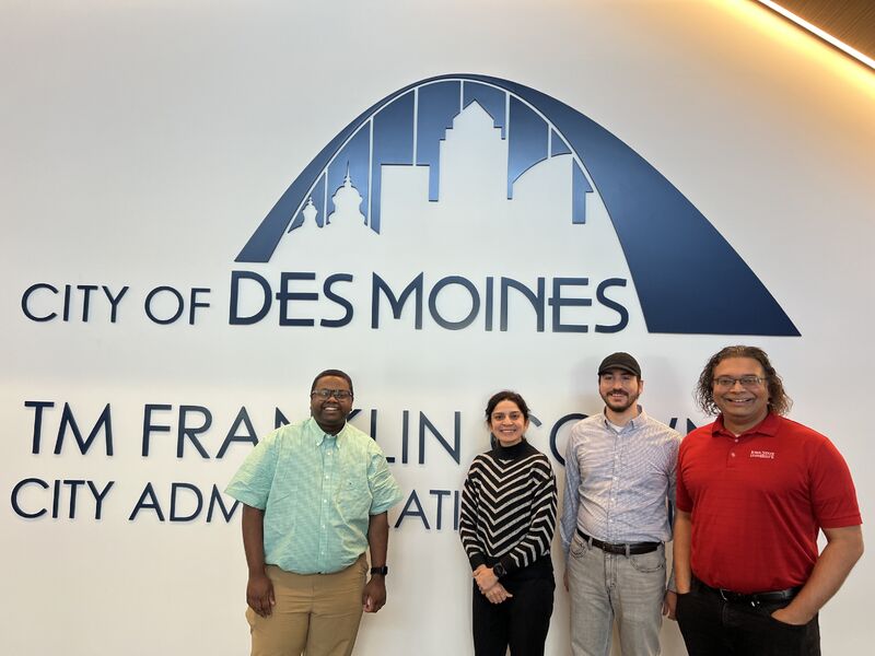 Four people standing in front of a City of Des Moines wall sign at the city administration office.