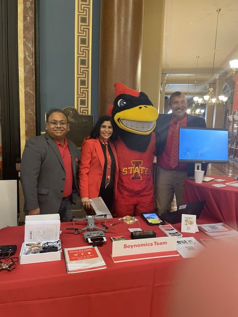 Three people and the Cy the Cardinal mascot behind a red-draped table with drones and a "Soynomics Team" sign in the Capitol.