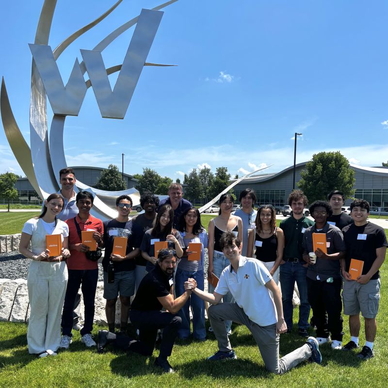 About twenty students and mentors holding orange folders in front of a large metal sculpture on a sunny lawn.