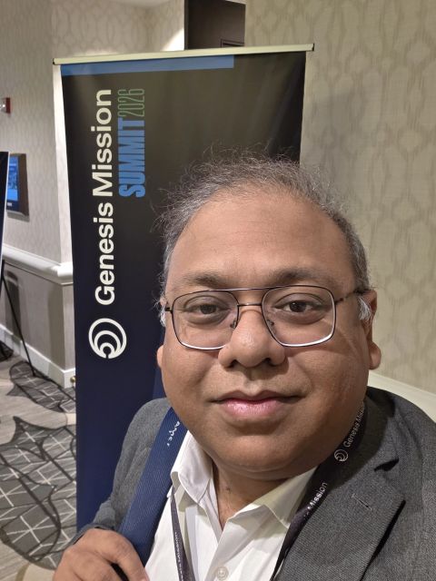 Selfie of a man in glasses in front of a Genesis Mission Summit 2026 banner.