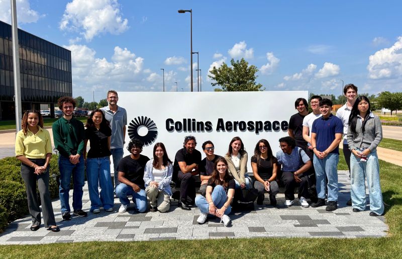 A group of about twenty students posing in front of a Collins Aerospace sign on a lawn.